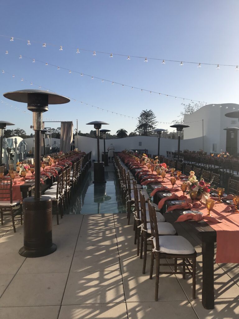Long outdoor dinner tables set on a terrace, with tall patio heaters along the tables and string lights hung overhead