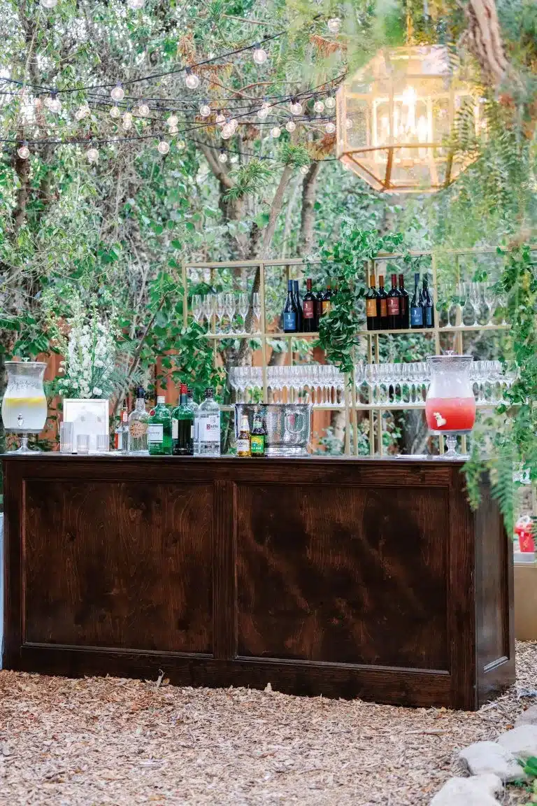 Stylish outdoor beverage station with wooden bar, assorted drinks, wine bottles, glasses, and decorative greenery, enhancing event ambiance and guest experience.
