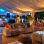 a sophisticated outdoor event setting is captured, showcasing a plush, elegantly designed tent with ambient lighting, perfectly balancing an open-air feel and an intimate atmosphere, surrounded by stylish decor and chic lounge furniture.