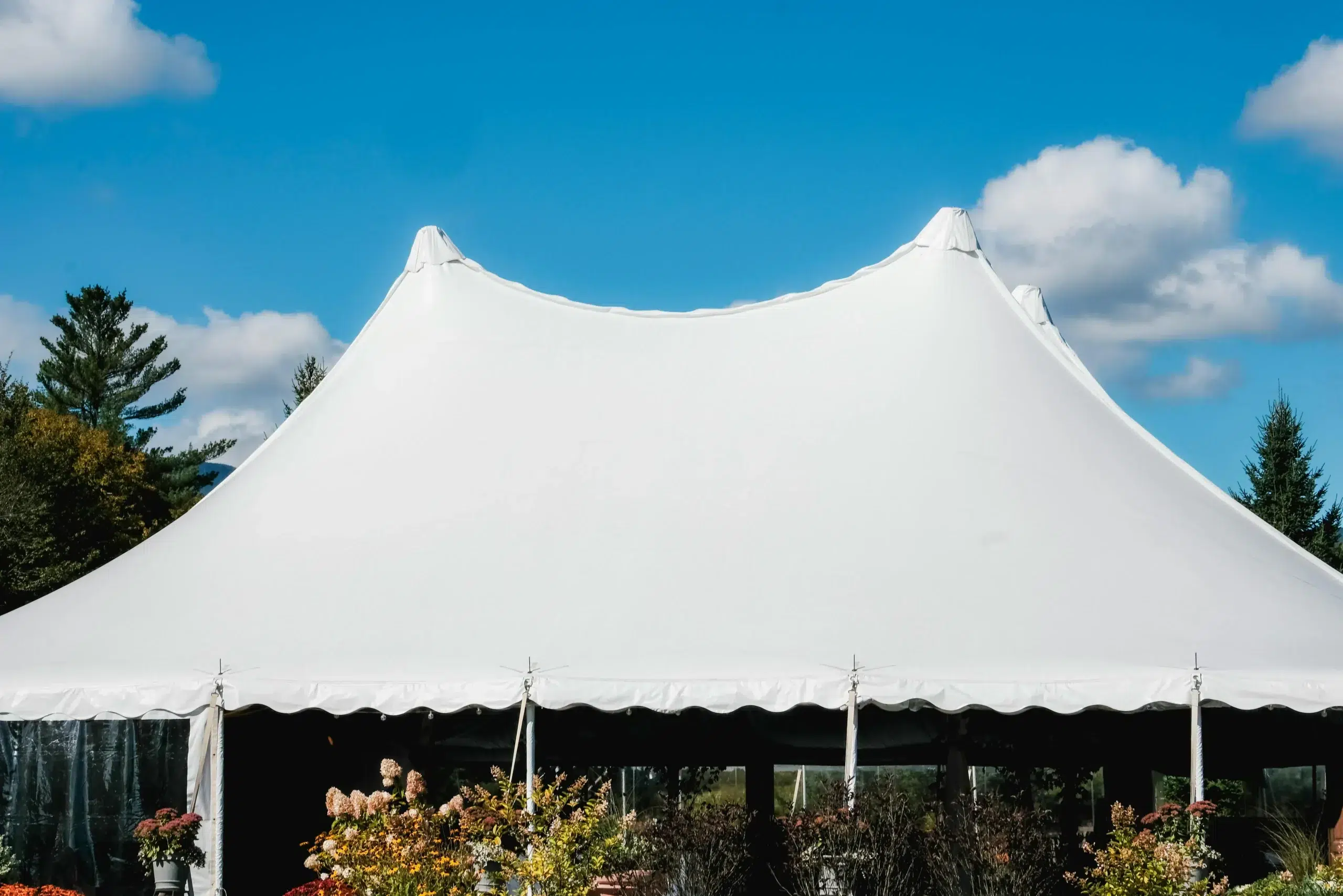 White frame tent with peaked design, set against a blue sky and surrounded by greenery, showcasing its unobstructed interior space ideal for events.