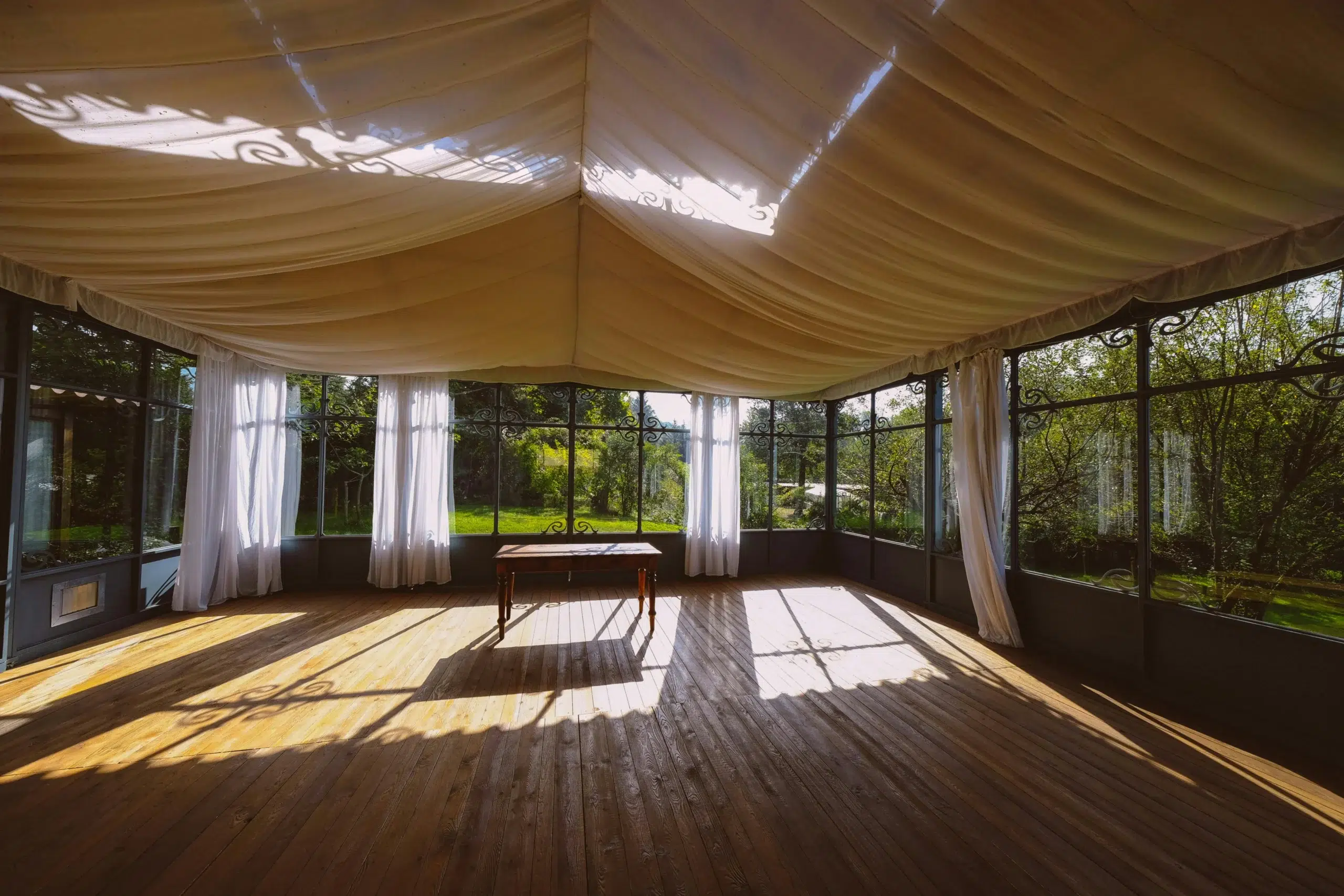 Interior of a tent with draped fabric ceiling, wooden floor, and large windows showcasing lush greenery, ideal for outdoor events and weddings.