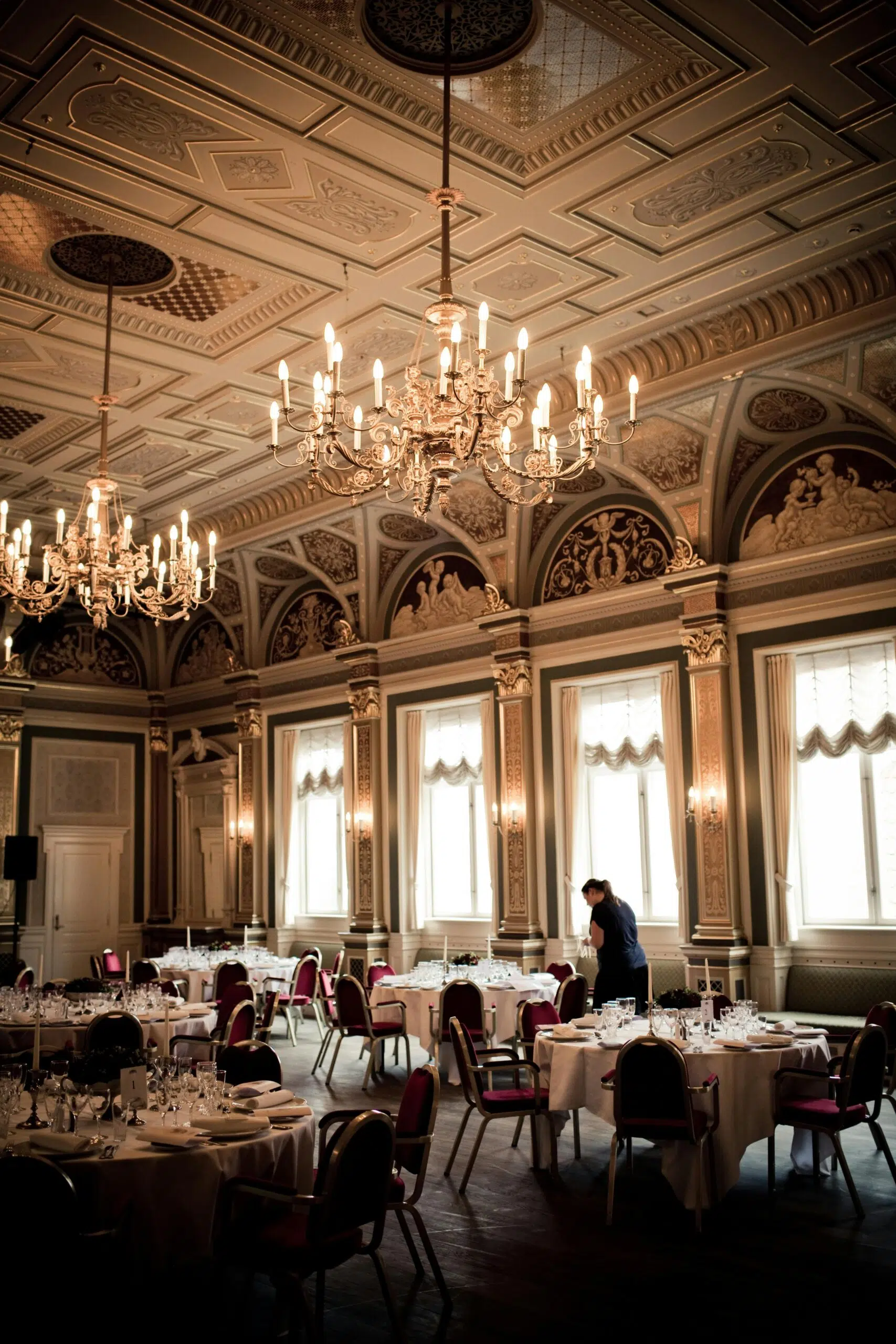 Elegant event space with chandeliers, ornate ceiling, and tables set for dining, showcasing potential for personalized decor and rental options.