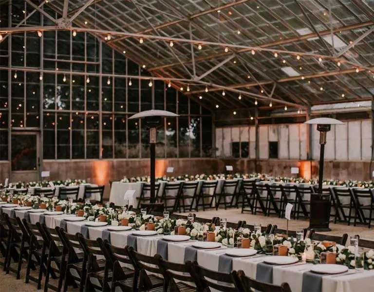 Industrial-style venue with long banquet tables, dark chairs, and hanging lights.