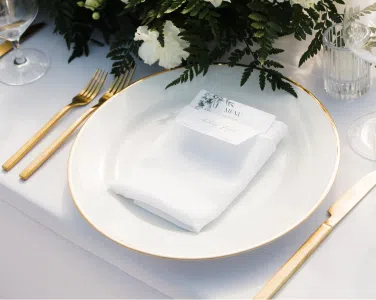 Elegant table setting featuring white china with gold rim, brushed gold flatware, and a neatly folded napkin, surrounded by greenery, reflecting rustic elegance for weddings at Tuscan Hills Estate.