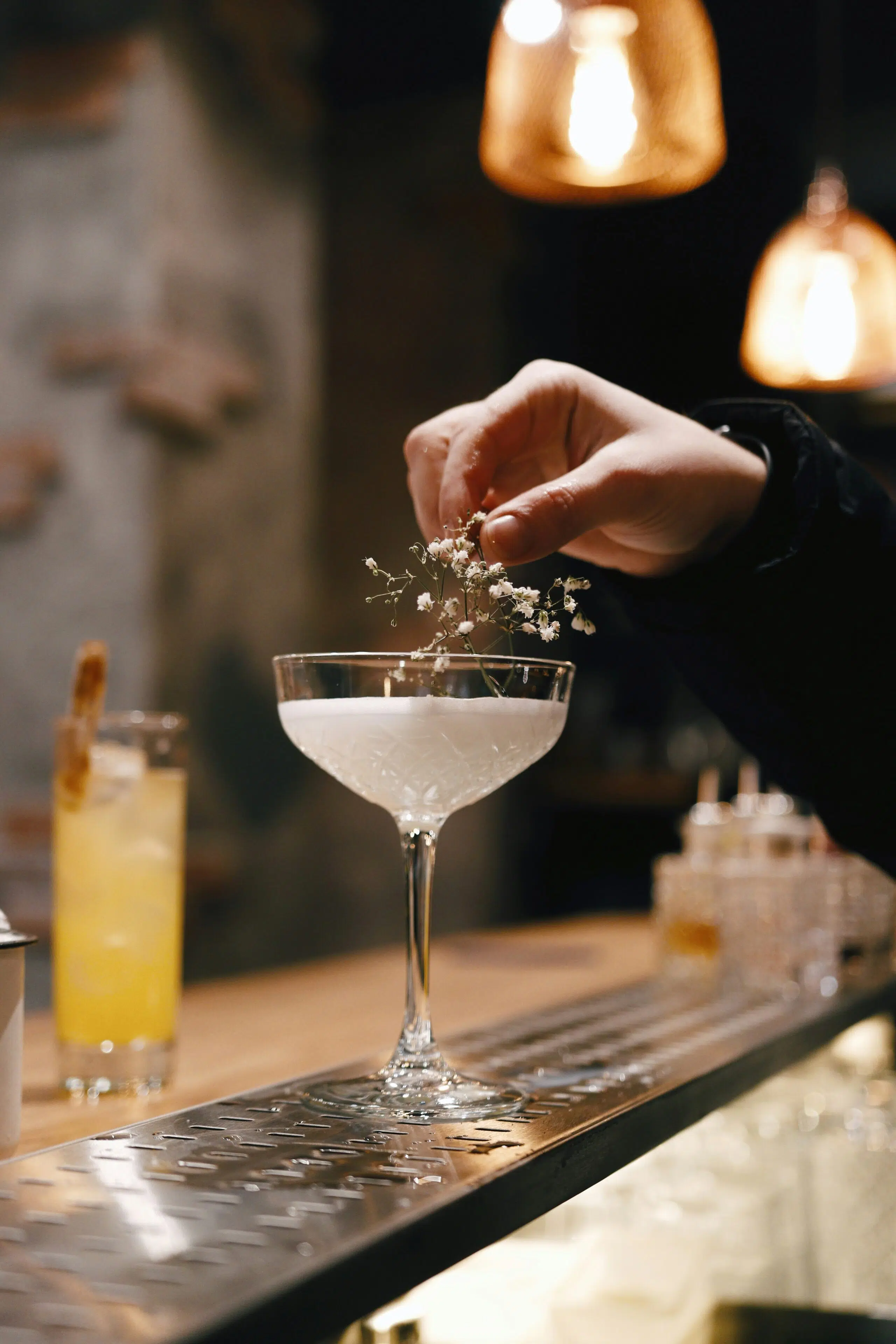 Hand placing delicate flowers on a cocktail in a stylish glass, with a blurred background of a bar featuring a yellow drink and decorative lighting, emphasizing elegant cocktail presentation for festive events.
