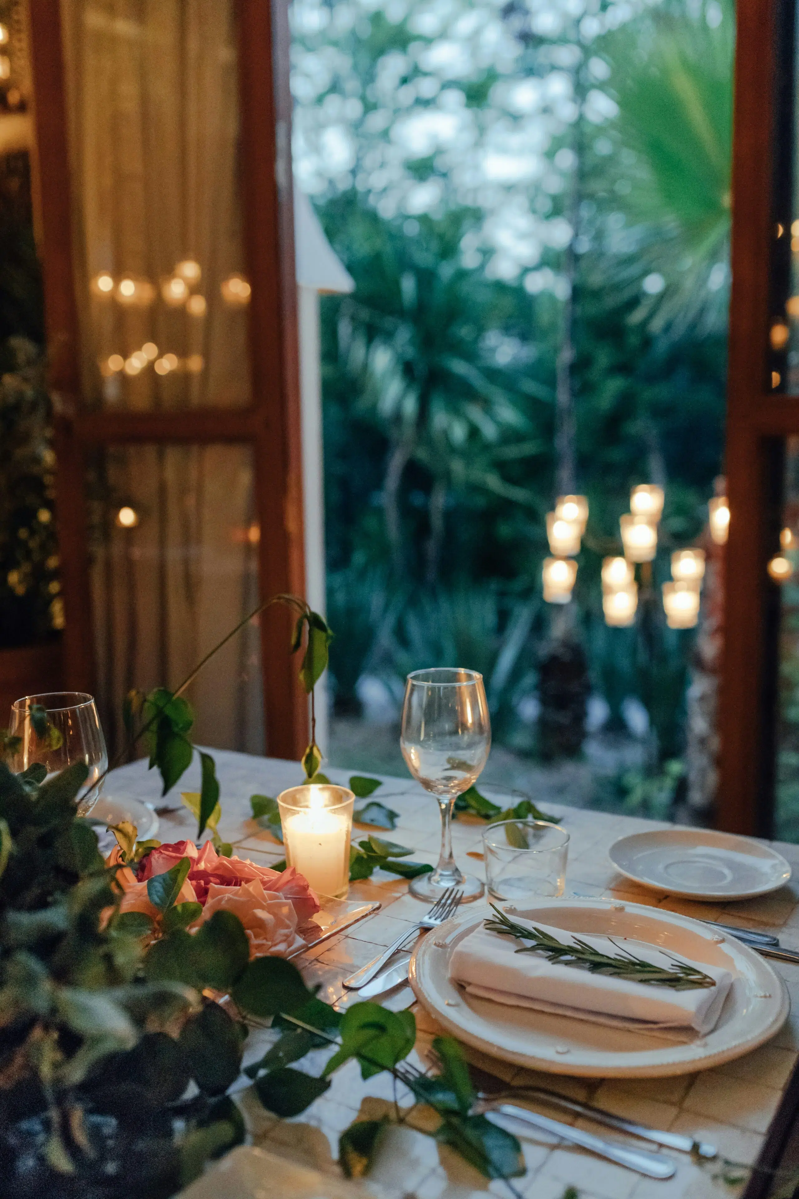 Elegant dining table set for a holiday celebration, featuring candles, glassware, fresh flowers, and greenery, with a backdrop of softly lit outdoor ambiance.