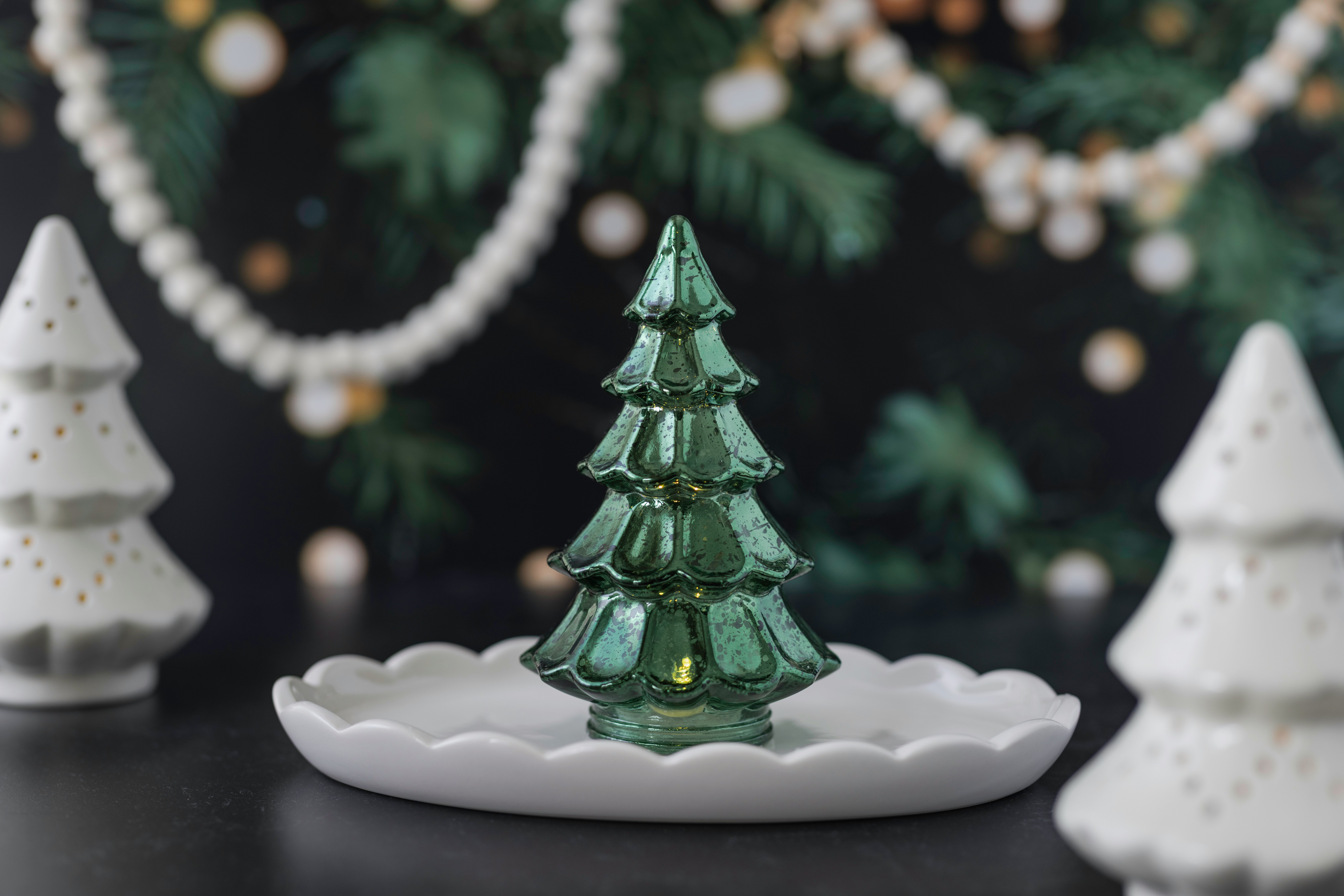 Green glass Christmas tree centerpiece with white ceramic trees and decorative garland in a festive setting.