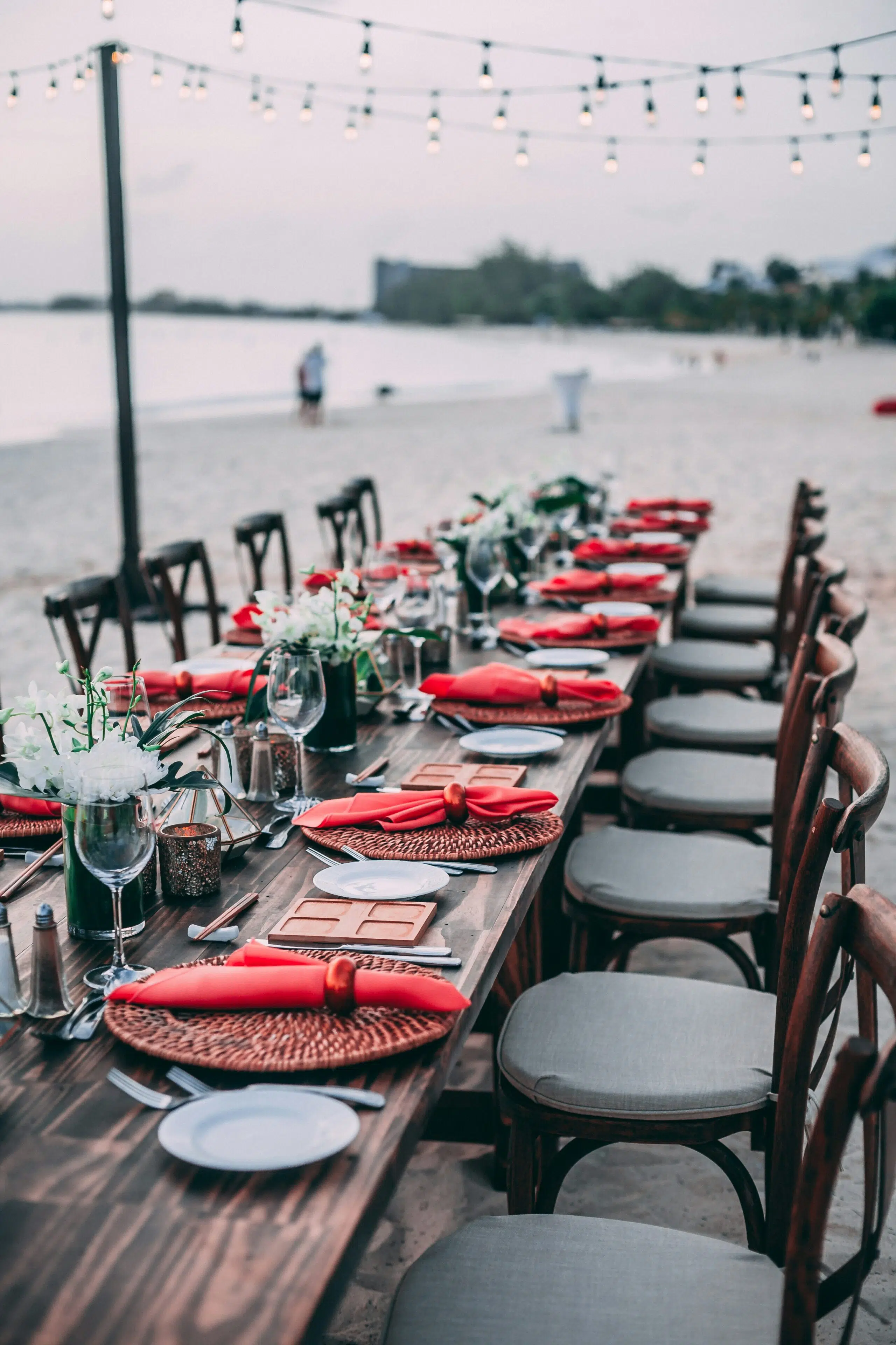 Beachfront dining setup with a long wooden table adorned with red napkins, woven placemats, glassware, and floral centerpieces, reflecting coastal wedding aesthetics.