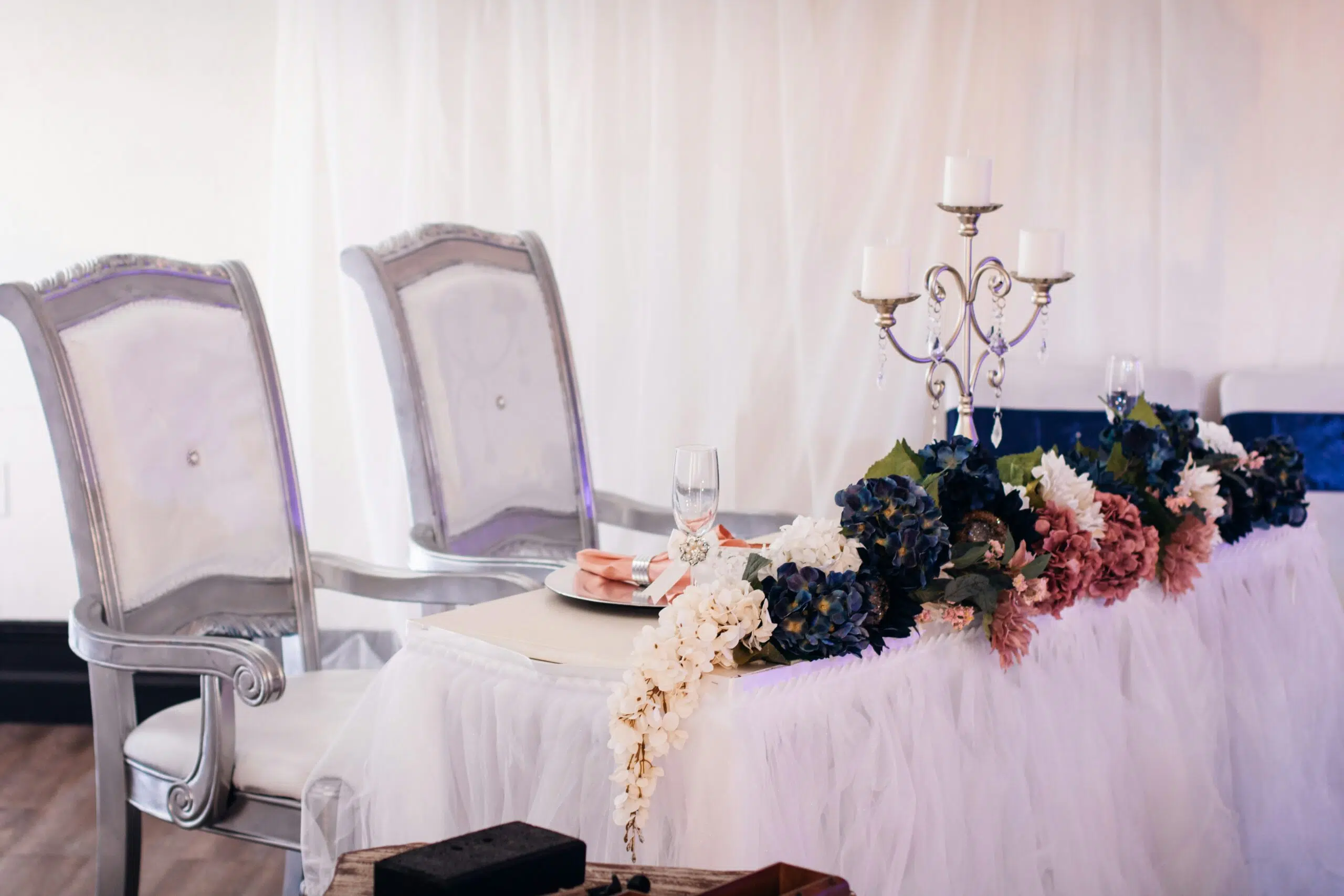 Elegant silver chairs with white upholstery beside a decorated table featuring floral arrangements, a silver candelabra, and dinnerware, set against a soft backdrop, suitable for formal events and gatherings.