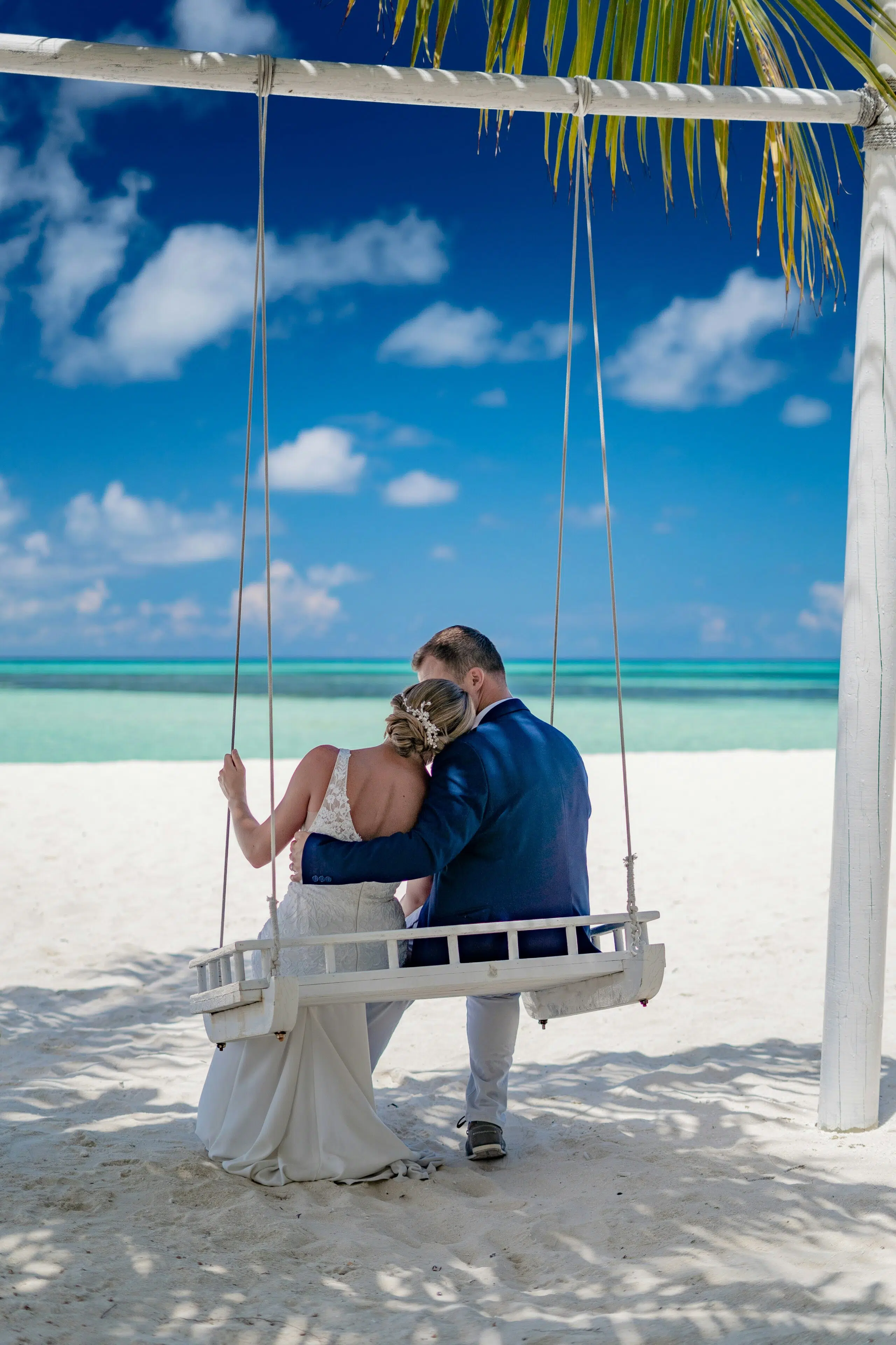 Couple sitting on a white swing at a beach, embracing with ocean and blue sky in the background, ideal for coastal wedding inspiration.