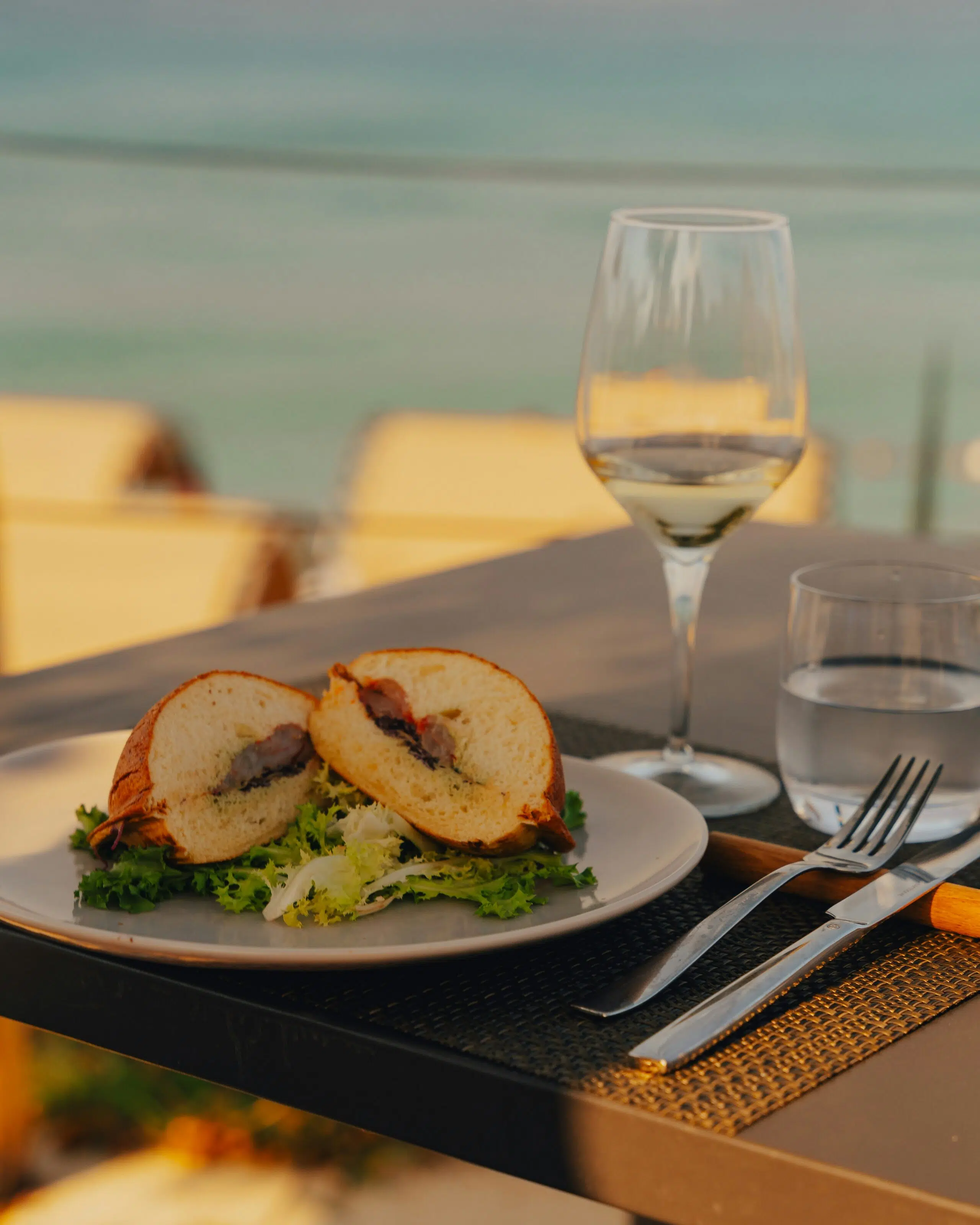 Plate of sandwich with lettuce, glass of white wine, and water, set against a coastal backdrop, emphasizing a relaxed dining experience for coastal weddings.