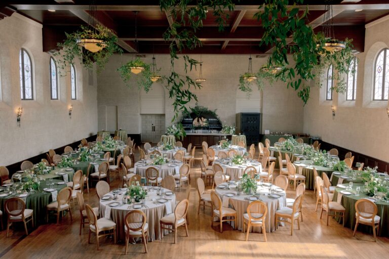 Elegant wedding reception setup featuring round tables adorned with greenery and floral centerpieces in a spacious venue with high ceilings and large windows, perfect for a romantic celebration.