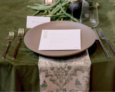 Elegant table setting featuring a round plate, menu card, Basic Matte Silver Flatware, and Verona Ridged Glassware on Olive Shantung Linen, enhancing the romantic atmosphere of a wedding reception at Hotel San Buena.