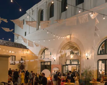 Wedding reception at Hotel San Buena, illuminated by bistro lighting, with guests mingling under decorative papel picado banners, showcasing an elegant outdoor ambiance.