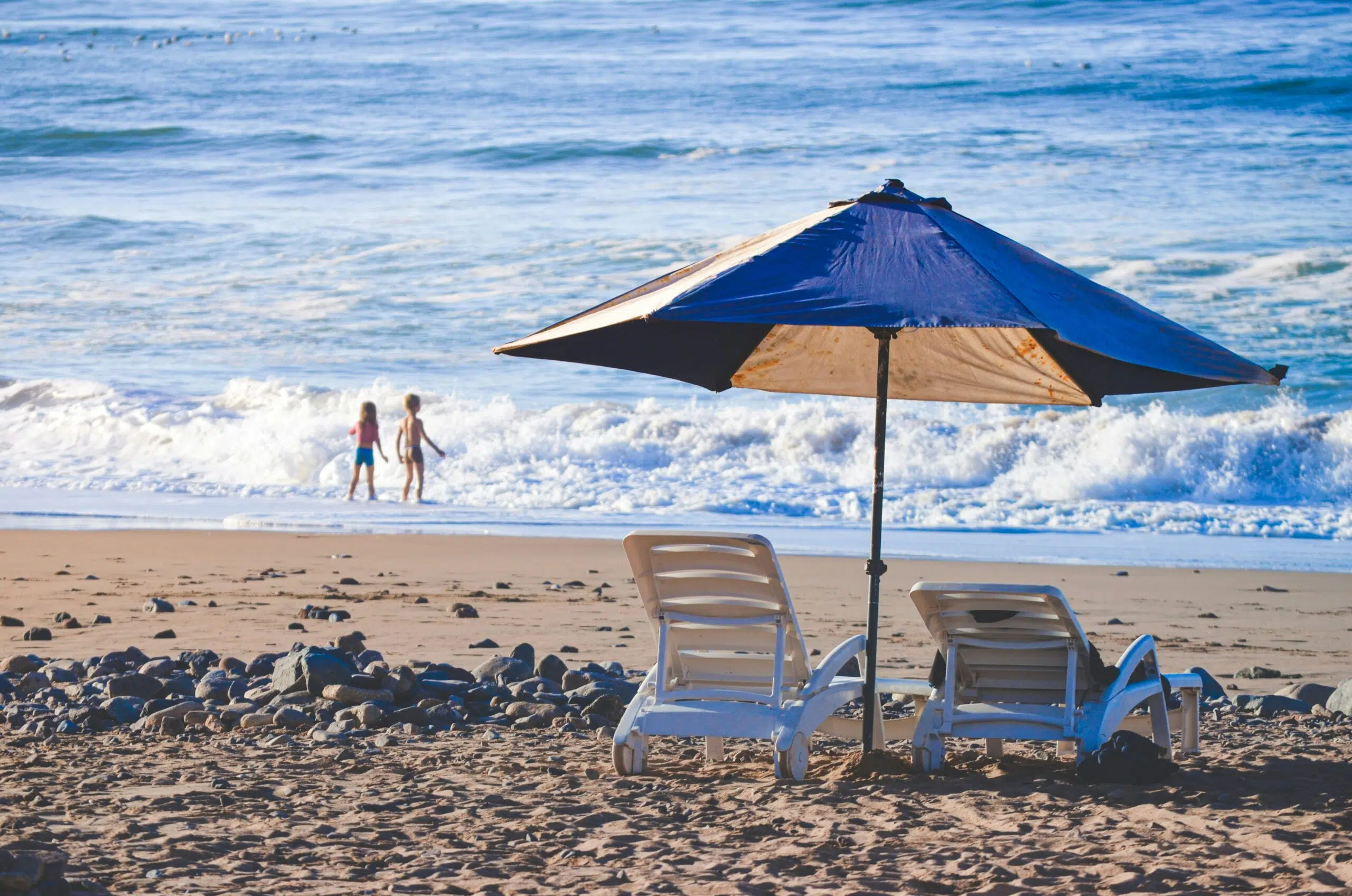 Beach scene with two white lounge chairs under a blue umbrella, overlooking children playing in the ocean waves, emphasizing outdoor event comfort in coastal environments.