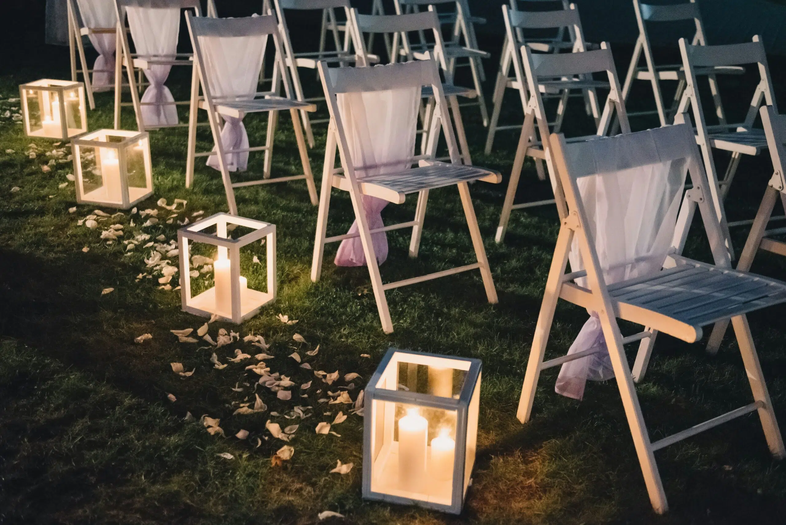 White folding chairs arranged for an outdoor event, illuminated by decorative lanterns and surrounded by rose petals, creating an inviting atmosphere for gatherings like weddings or parties.