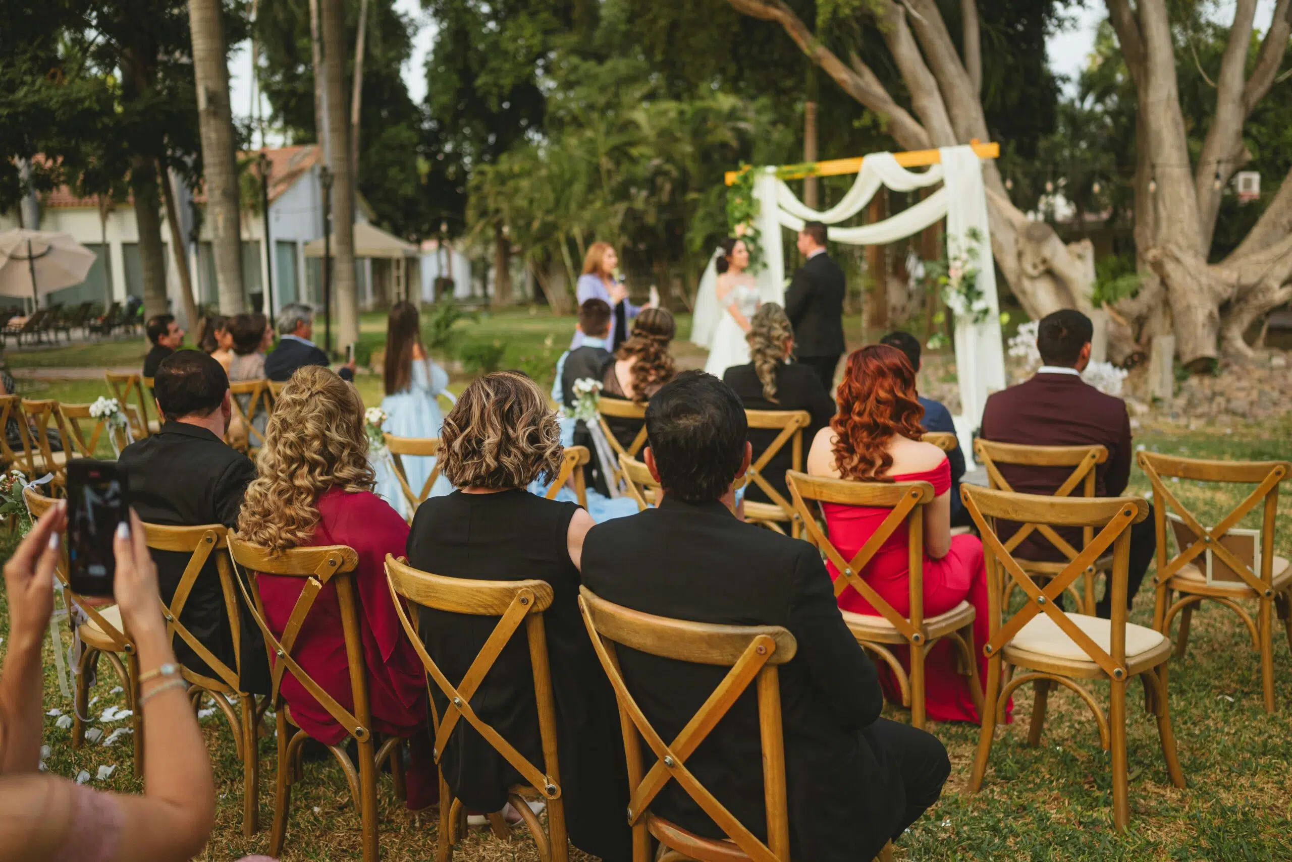 Outdoor wedding ceremony with guests seated on wooden chairs, bride and groom at altar with draped fabric and greenery, surrounded by trees in Ventura County.