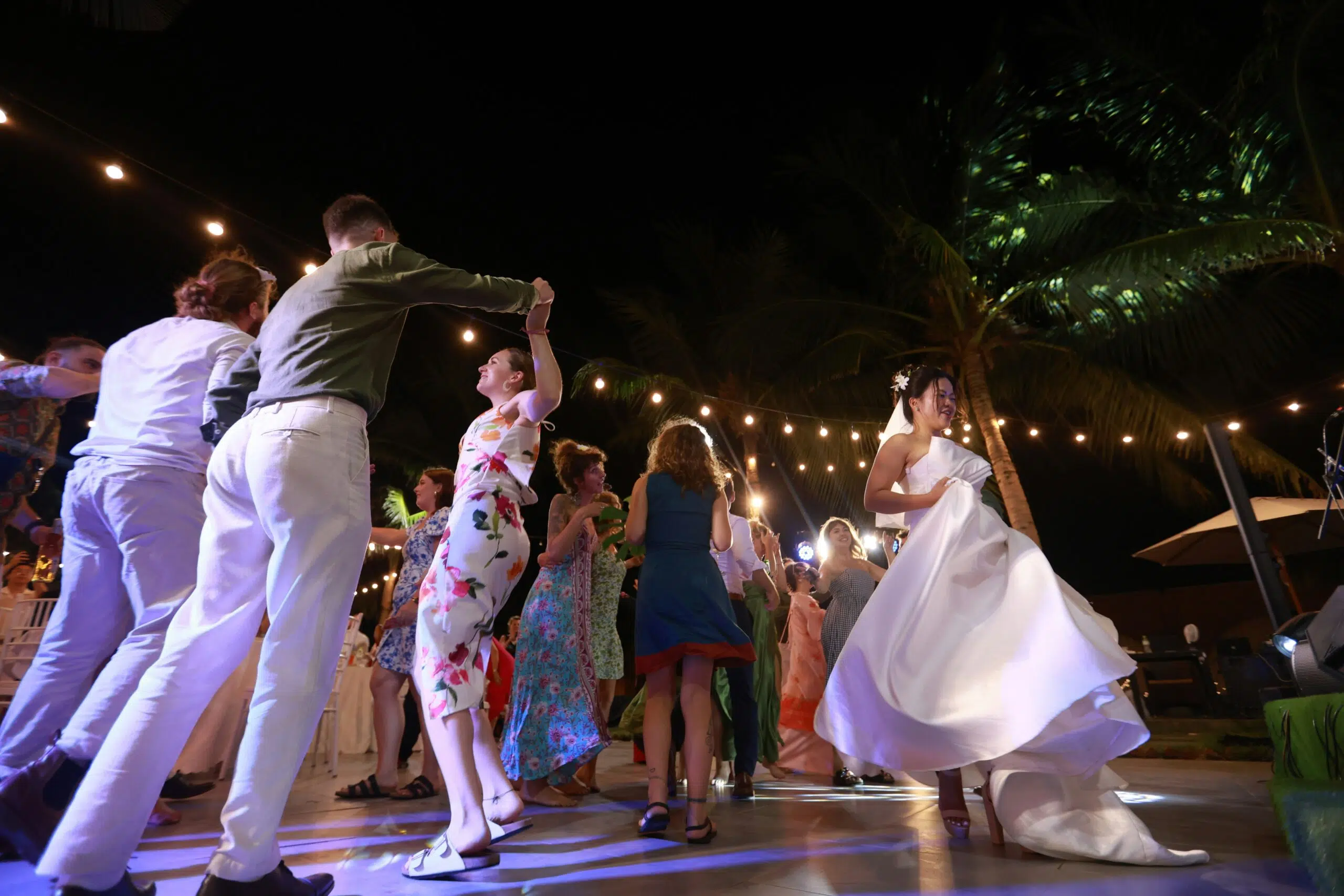 Couples dancing joyfully on a wooden dance floor at a wedding reception, surrounded by festive lighting and tropical palm trees, showcasing a lively celebration atmosphere.