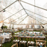 ### Key Elements
- **Setting**: Outdoor tented venue
- **Arrangement**: Multiple tables with seating
- **Decor**: Elegant floral centerpieces, blue table settings
- **Lighting**: Soft ambient lighting, possibly from hanging fixtures
- **Mood**: Festive and inviting atmosphere, suitable for a celebration or event
The image depicts a beautifully arranged outdoor tent, filled with tables set for a gathering. Each table is adorned with floral centerpieces and blue dinnerware, creating a cohesive and elegant look. The soft lighting enhances the inviting ambiance, suggesting a celebratory theme, likely for a wedding or special occasion. The overall mood is warm and welcoming, making it an ideal setting for socializing and celebration.