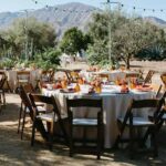 ### Setting
- Outdoor venue with mountains in the background.
- Natural landscape featuring trees and agave plants.
### Table Arrangements
- Round tables set with beige tablecloths.
- Wooden chairs surrounding the tables.
### Decorative Elements
- Centerpieces with small floral arrangements.
- Orange glassware and plates.
### Lighting
- Strings of lights adding a festive ambiance.
### Overall Mood
The atmosphere conveys a rustic and inviting feel, ideal for a casual yet elegant gathering. The natural setting enhances a sense of warmth and celebration.