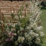 ### Key Elements
- **Main Subjects**: Floral arrangements
- **Objects**: Wooden chairs, greenery
- **Notable Features**: Lush, diverse flowers; organized seating
- **Text/Symbols**: None present
### Overall Mood/Theme
The image conveys a serene and romantic atmosphere, typical of a wedding or outdoor event setting. The abundance of white and pastel flowers creates a soft, elegant aesthetic, while the neatly arranged chairs suggest preparation for a gathering, enhancing the sense of anticipation and celebration.