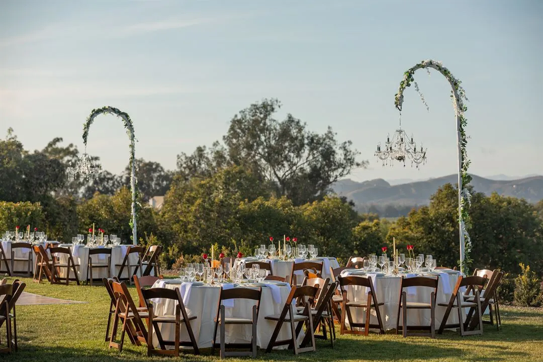 Outdoor Santa Barbara reception with round dining tables, wood folding chairs, floral chandeliers, and mountain views.