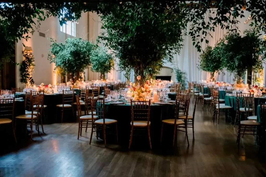 Elegant indoor fall reception with dark dining tables, wood chairs, tall trees, floral centerpieces, and warm ambient lighting.