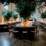 Elegant indoor fall reception with dark dining tables, wood chairs, tall trees, floral centerpieces, and warm ambient lighting.