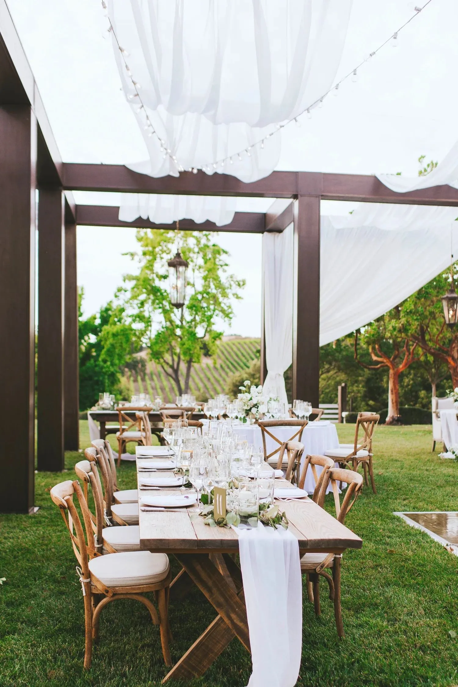Long wood dining table with cross-back chairs, white flowers, glassware, and overhead draping at an outdoor vineyard event.