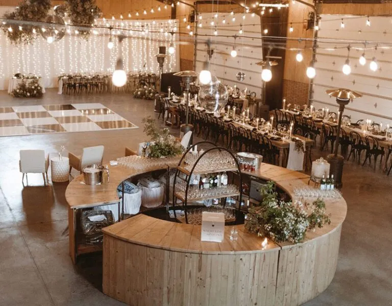 Large circular wood bar surrounded by reception tables, string lights, lounge seating, and a dance floor inside an event venue.