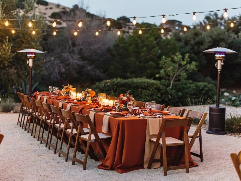 Long outdoor fall dinner table with wood folding chairs, rust-colored linens, candlelight, string lights, and patio heaters.